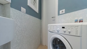 A modern laundry room with a front-loading washing machine, a tiled wall, and a door leading to another room. - Italianway - Bezzecca 12 (Milan)