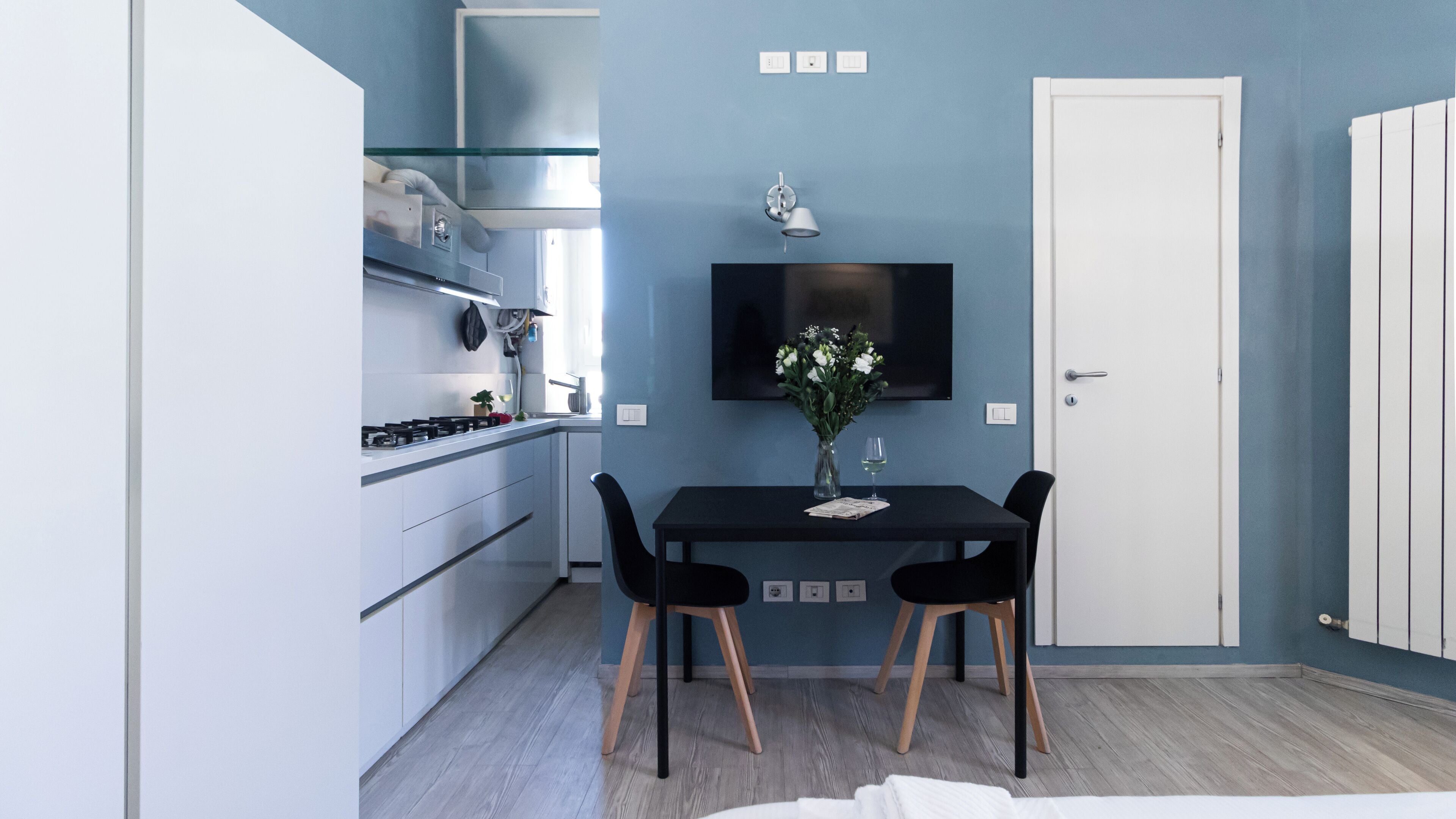 A compact kitchen with modern appliances, a dining table with chairs, and a flat-screen TV mounted on the wall.