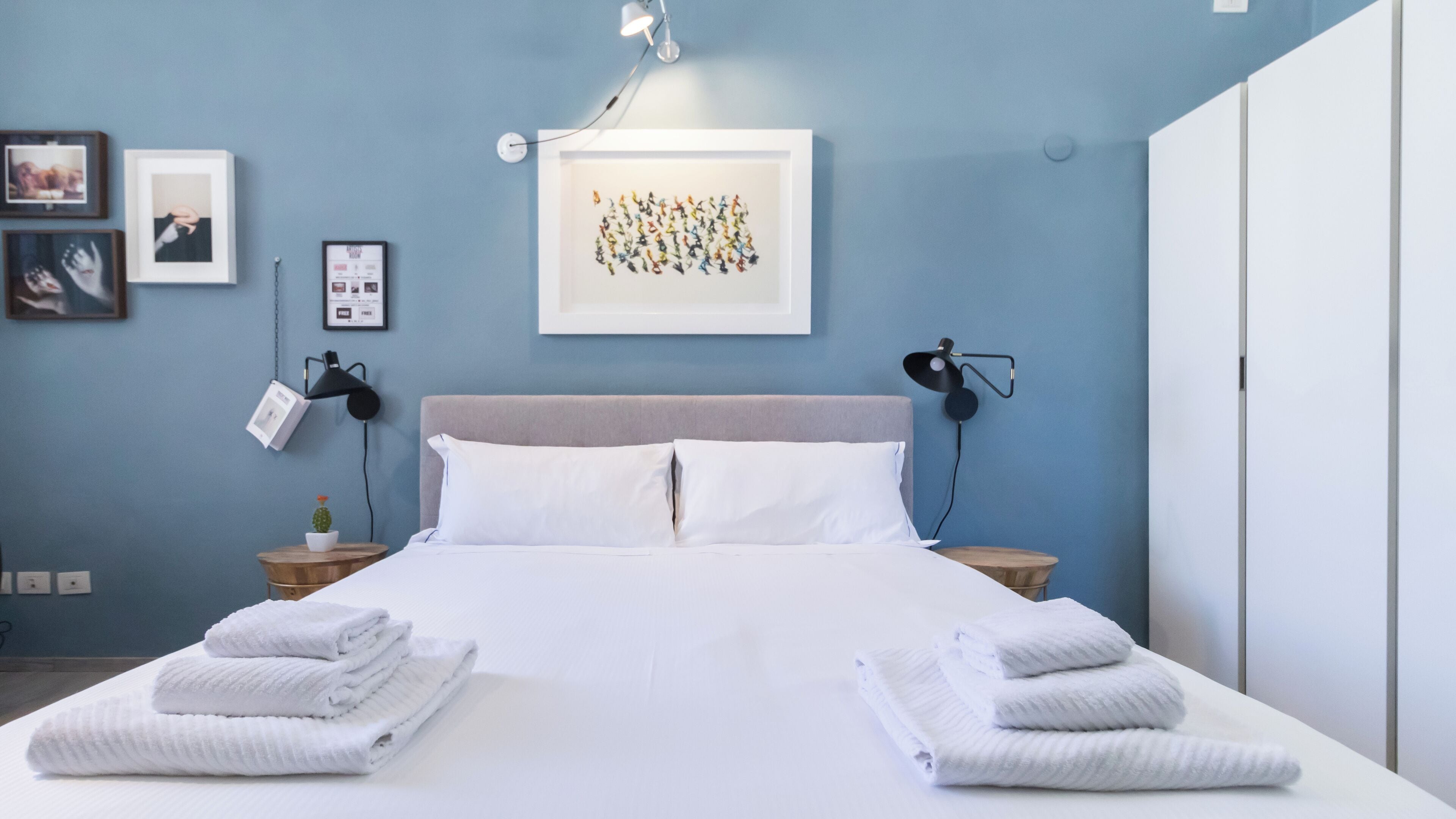 A neatly made bed with white linens and folded towels, a headboard, bedside lamps, and a framed artwork on the wall.