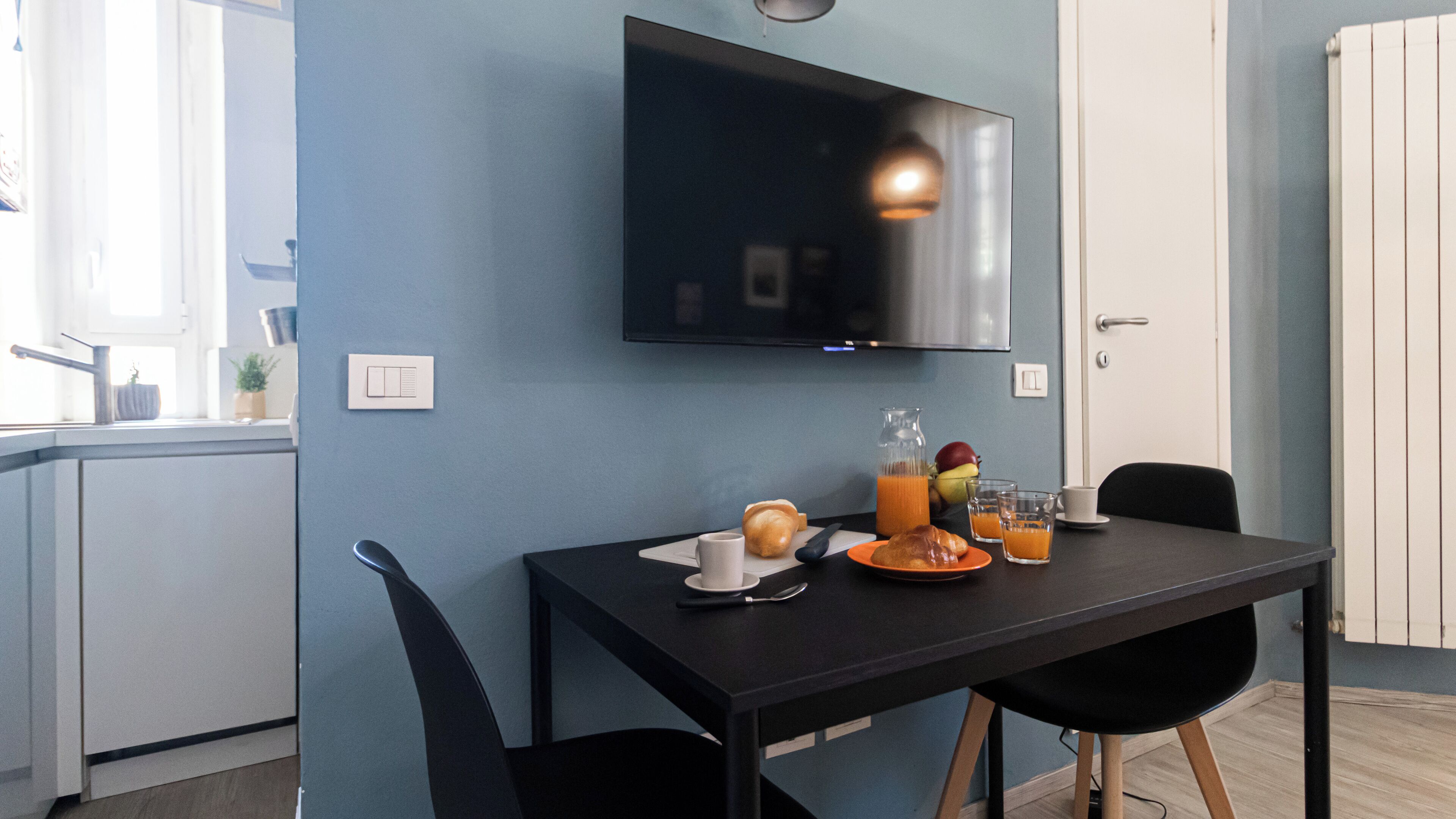 Une cuisine moderne avec un téléviseur à écran plat fixé au-dessus du comptoir, une table à manger avec des chaises, et un coin petit-déjeuner avec des croissants, des fruits et du jus d’orange.