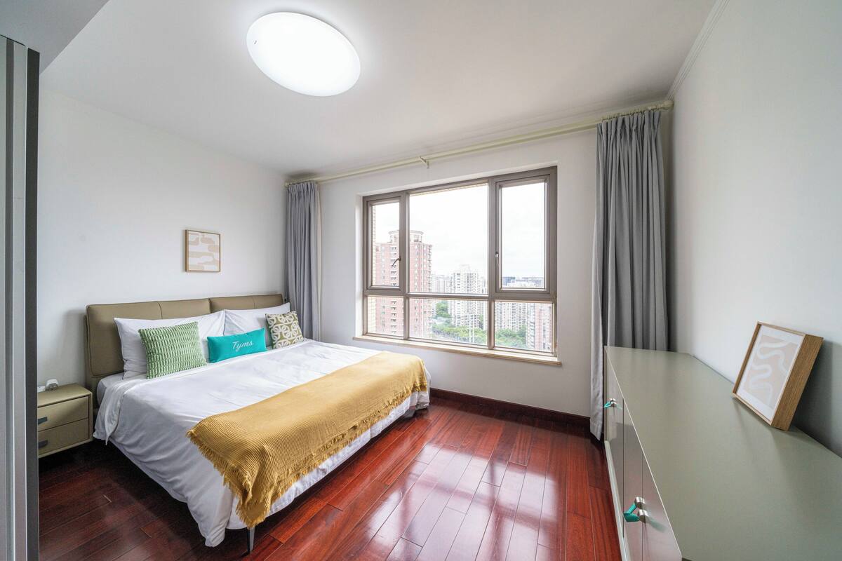 Un dormitorio con cama, mesita de noche, cómoda, ventana con cortinas y vista a la ciudad.