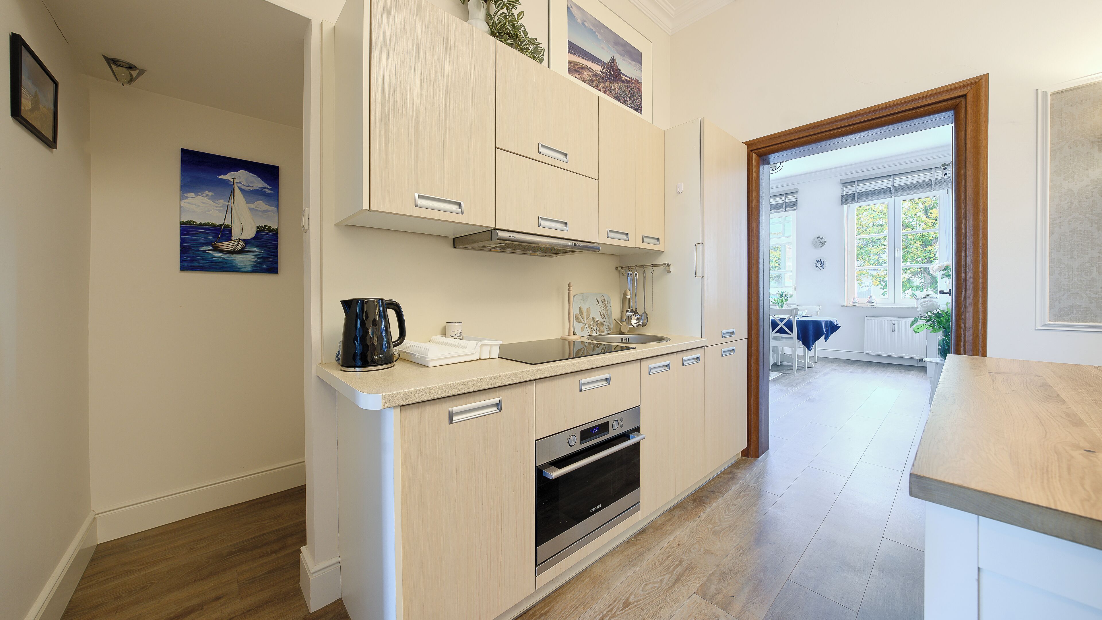 A modern kitchen with a built-in oven, a kettle, and a sink. There is a dining area visible through an open door.