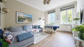 A living room with a grey sofa, a dining table with chairs, a white radiator, and a window with a view of greenery. - Apartamenty Sun & Snow Pomorska Przystań (Kołobrzeg)