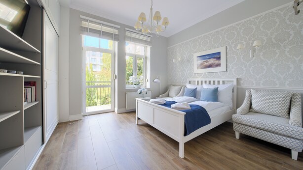 A bedroom with a bed, a chair, a nightstand, a window with blinds, and a bookshelf. - Apartamenty Sun & Snow Pomorska Przystań (Kołobrzeg)