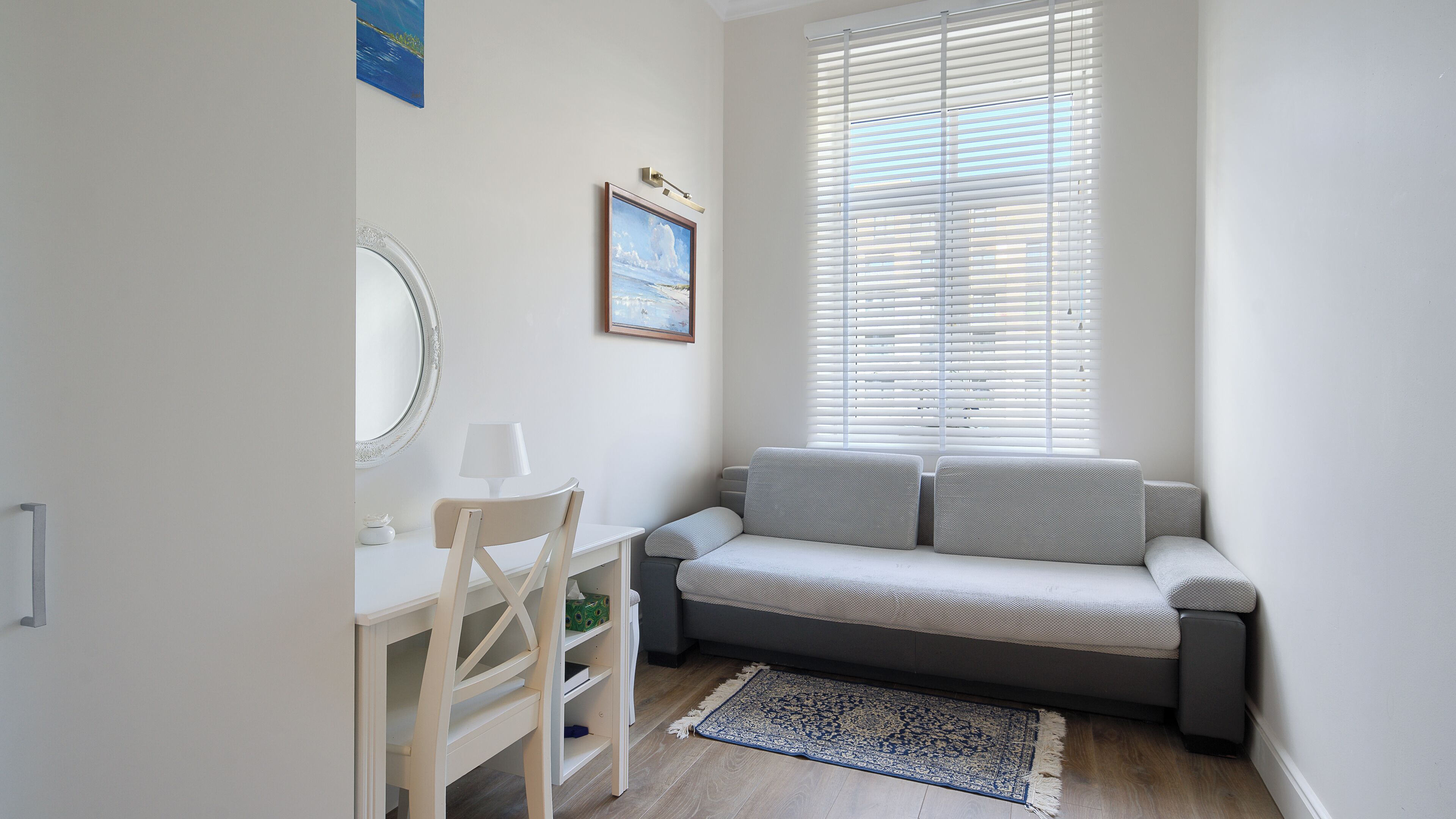 A room with a white desk, a chair, a grey sofa, a patterned rug, and a large window with blinds.