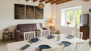 Un coin repas avec une table dressée pour quatre personnes, un canapé, un téléviseur et une fenêtre donnant sur un paysage verdoyant.