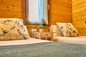 A wooden cabin room with two beds, a small table with a lamp, and a window with curtains.