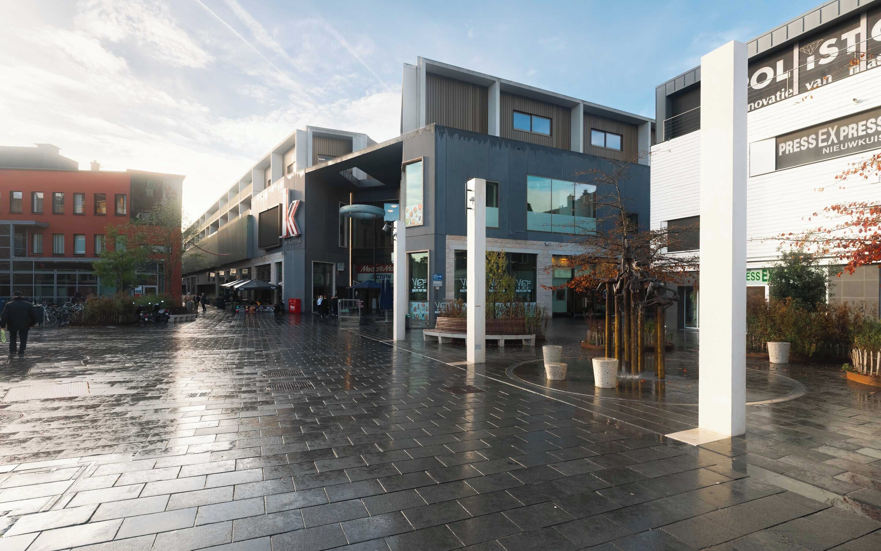 A modern urban plaza with a wet pavement, surrounded by contemporary buildings with signage for 'K' and 'Press Ex Press'.