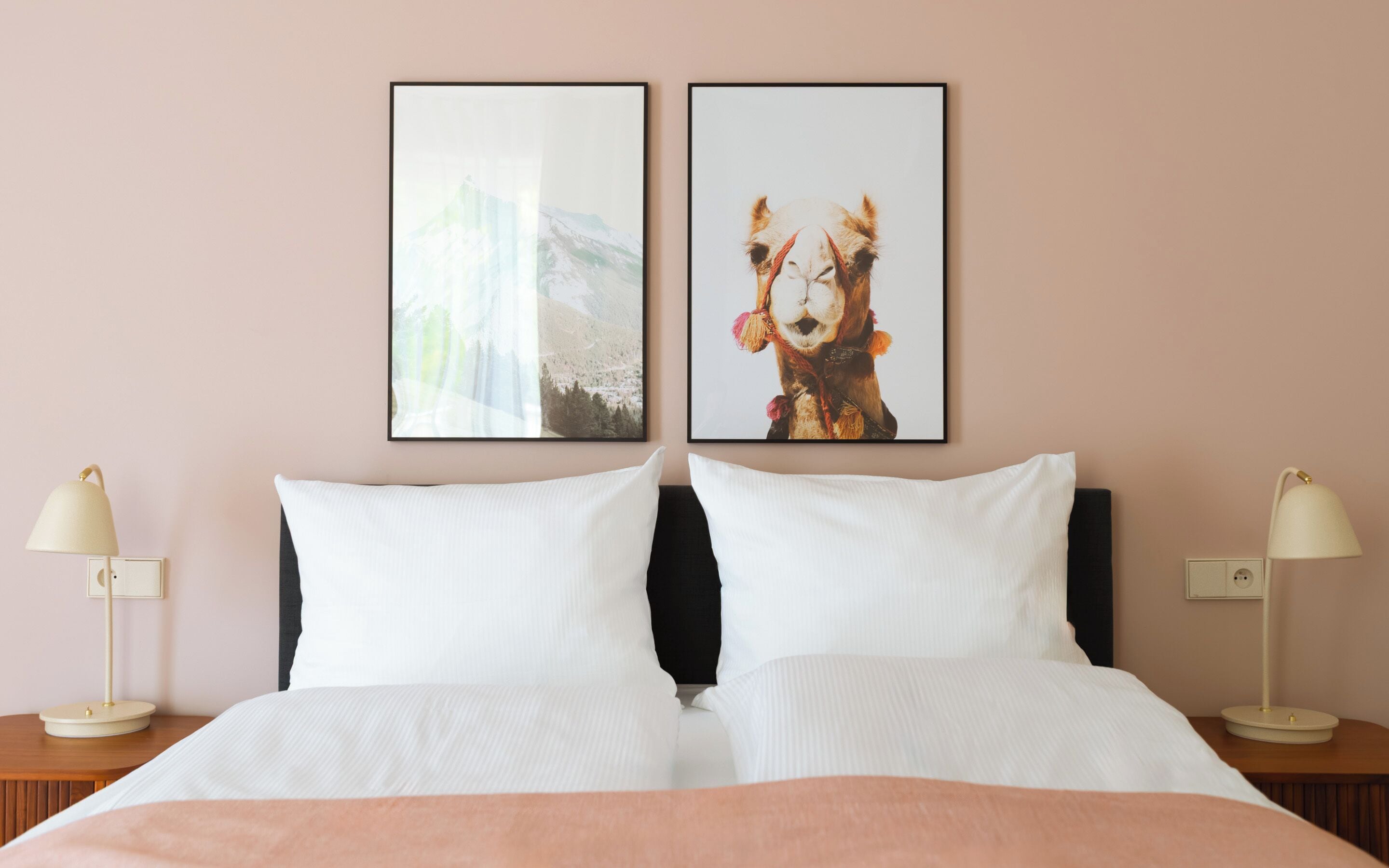 Cama bem arrumada com roupa de cama branca, duas almofadas e um cobertor cor de pêssego. Há duas mesas de cabeceira com luminárias e quadros nas paredes acima da cama.