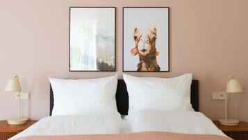 Cama bem arrumada com roupa de cama branca, duas almofadas e um cobertor cor de pêssego. Há duas mesas de cabeceira com luminárias e quadros nas paredes acima da cama.