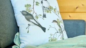 A grey sofa with a light blue pillow featuring birds and leaves, a light blue throw blanket, and a magazine on the table.