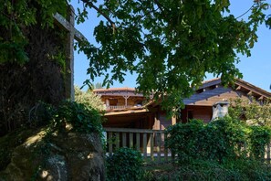 A rustic wooden cabin with a balcony, surrounded by lush greenery and a stone wall.