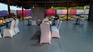 An outdoor dining area with white chairs and tables, a thatched roof, and a view of a colorful fence. - Wayma Paradise Resort (Marumbi)