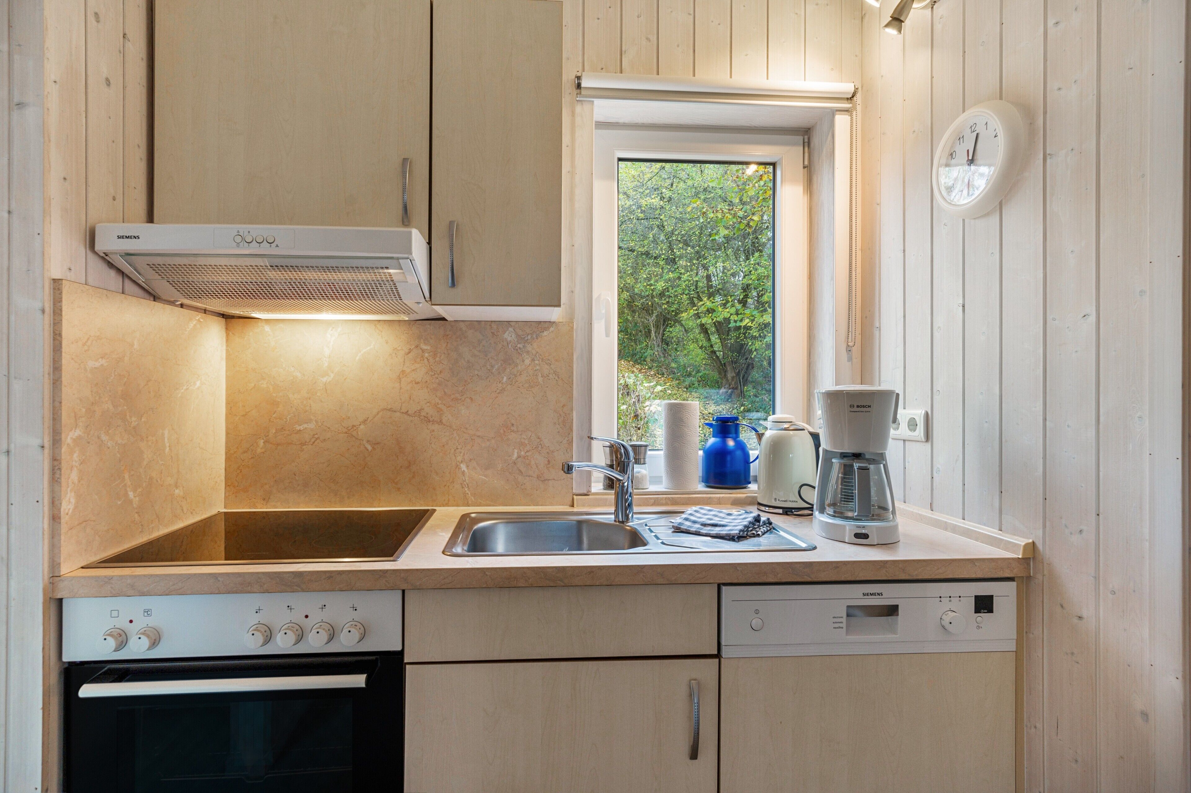 Una cocina con estufa Siemens, un fregadero, una ventana con vista a árboles y un reloj en la pared.
