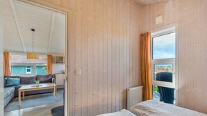 A room with wooden walls, a bed with patterned bedding, a window with curtains, and a view of an outdoor area with a sofa and table. - Holiday Home 'Skipper-Lodge West' with Sea View, Private Terrace and Wi-Fi (Kosel)