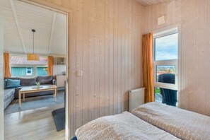 A room with wooden walls, a bed with patterned bedding, a window with curtains, and a view of an outdoor area with a sofa and table. - Holiday Home 'Skipper-Lodge West' with Sea View, Private Terrace and Wi-Fi (Kosel)