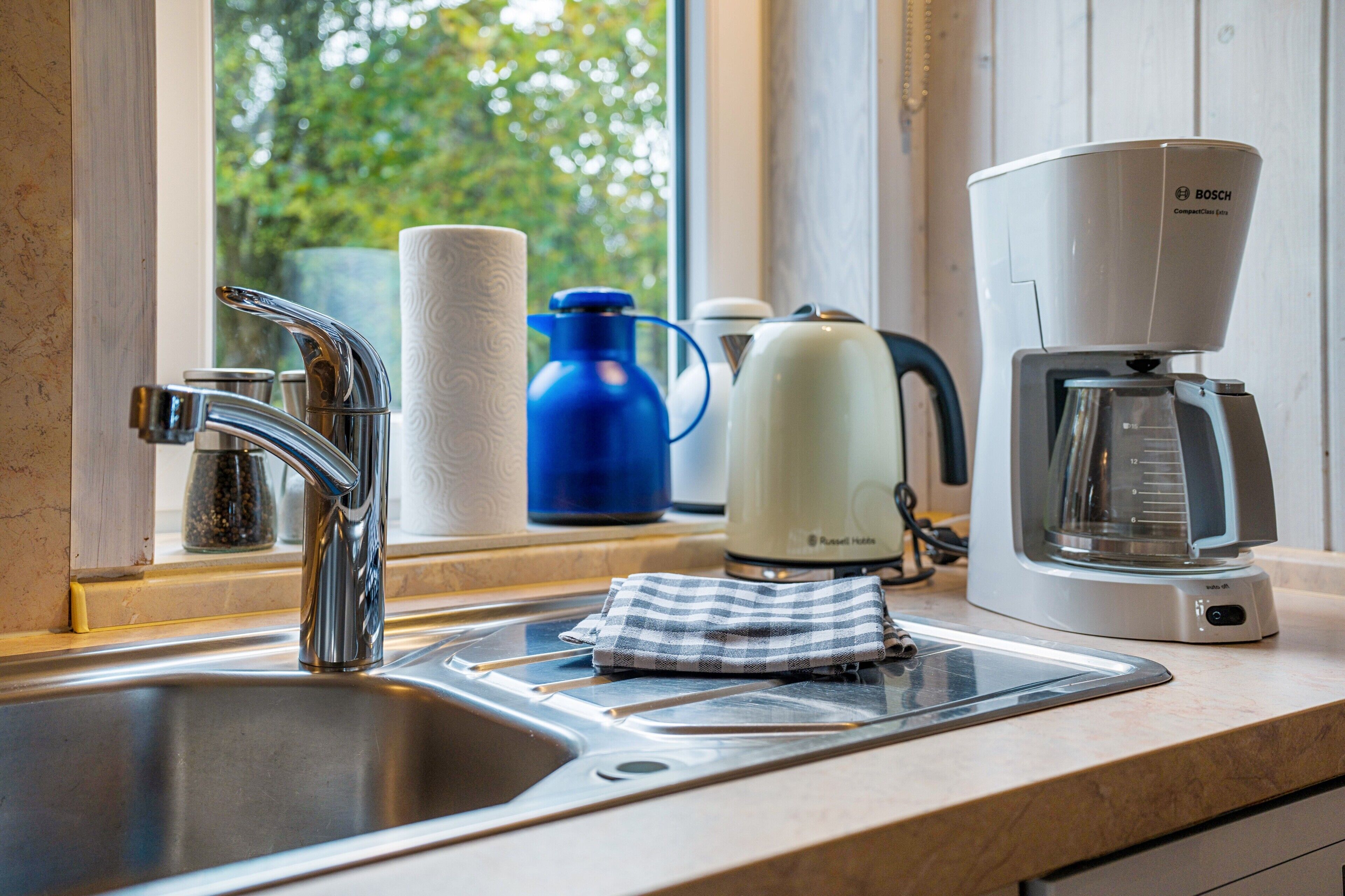 A kitchen with a stainless steel sink, a Bosch coffee maker, a blue water bottle, and a white kettle.