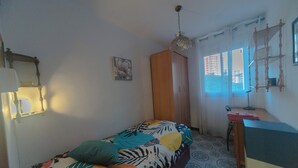 A small, simple room with a bed, a wooden wardrobe, a desk, and a window with curtains.