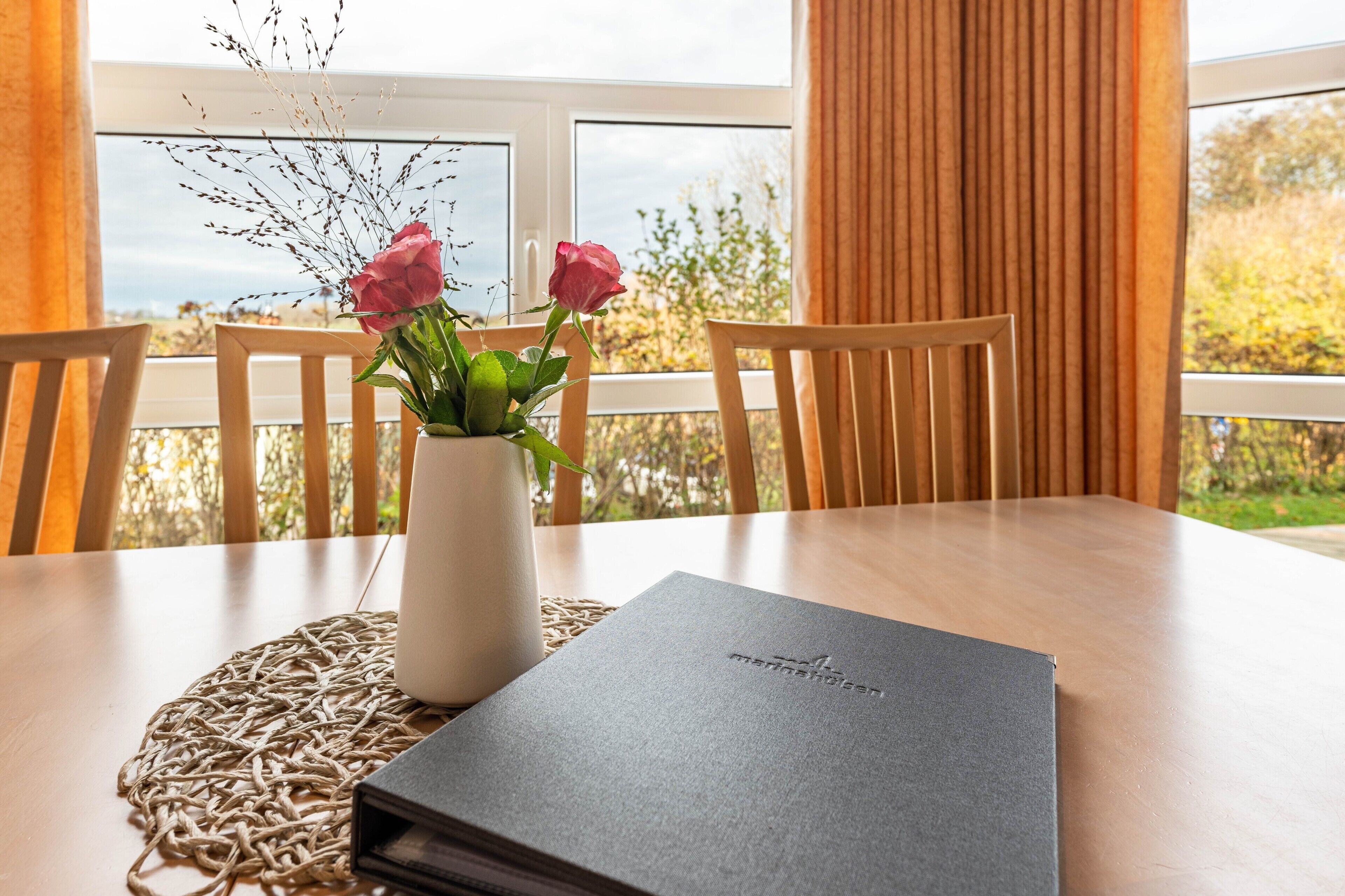 Une table à manger ornée d’un vase de fleurs, d’un livre, et offrant une vue sur la verdure par la fenêtre.