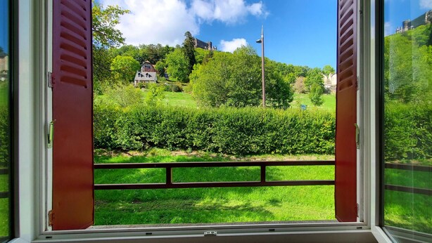 A window with a view of a lush green landscape, a house, and a hill with a castle-like structure in the background.