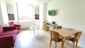 A well-lit room with a red sofa, a dining table with chairs, a TV mounted on the wall, and a window with curtains.