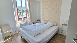 A bedroom with a bed, a chair, a window with a view of houses, and a radiator.