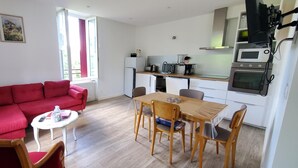 Un espacio habitable compacto con un sofá rojo, una pequeña mesa de comedor con sillas, una kitchenette con microondas y horno, y una ventana con cortinas.