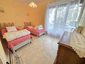 A room with two beds, a wooden desk, and a window with sheer curtains.