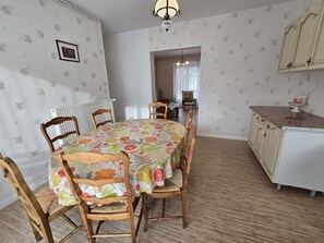 A dining area with a round table covered in a floral tablecloth, surrounded by wooden chairs with woven seats. A white radiator is visible on the left wall, and a wooden cabinet with a countertop is on the right.
