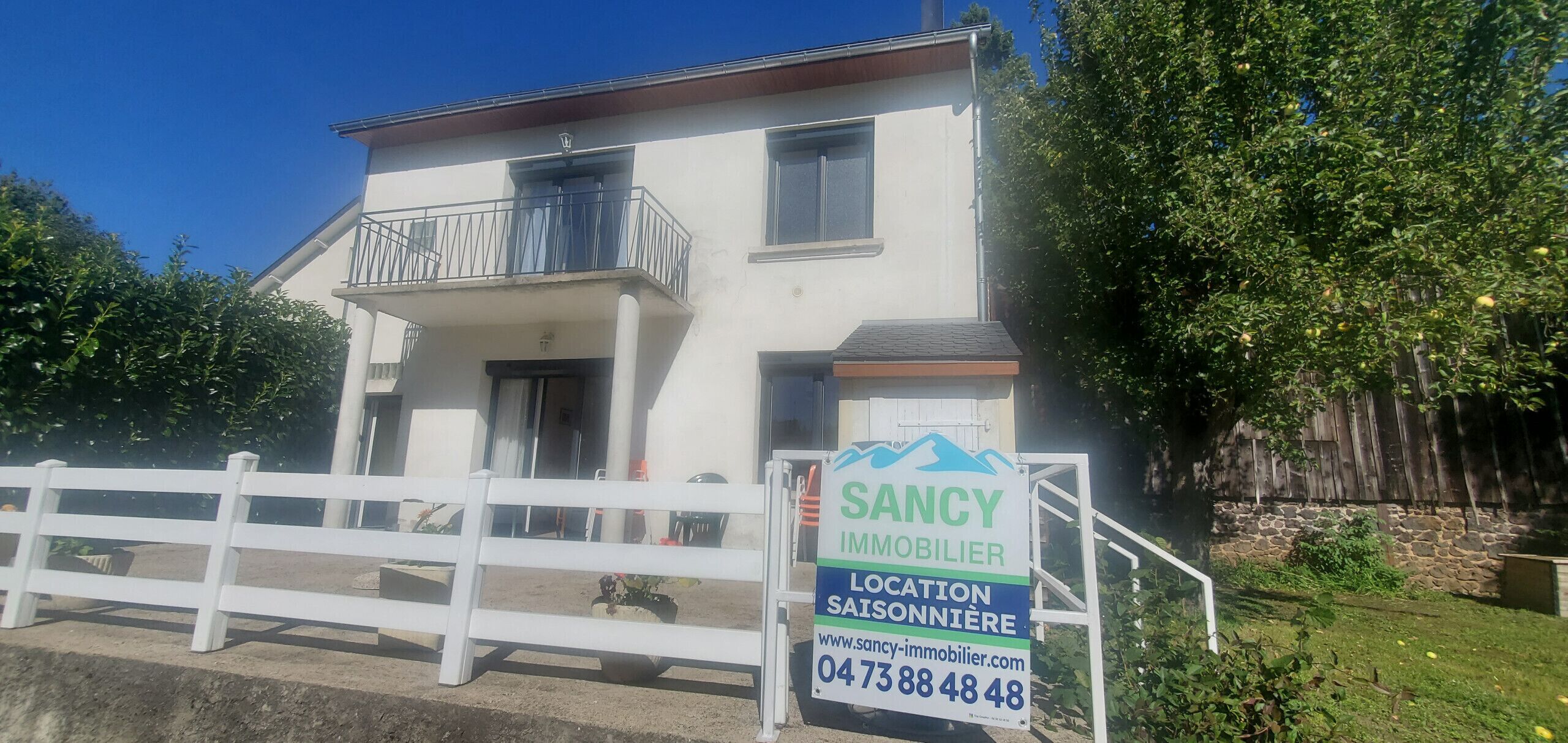 Une maison à deux étages avec un balcon, une clôture blanche à l’avant et une enseigne publicitaire pour des services immobiliers.