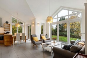 A spacious living area with a dining table, two armchairs, a sofa, and large windows offering a view of a garden.
