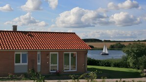 Uma casa com telhado de telhas vermelhas, o número '28' visível e vista para um lago com um veleiro.