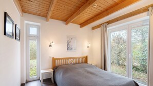 Um quarto com teto de madeira, uma cama com cobertor cinza, duas mesas de cabeceira, uma janela com cortinas e um quadro na parede.