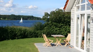 Uma casa com vista para um lago e um veleiro, um pátio com cadeiras e uma mesa, e um telhado vermelho.