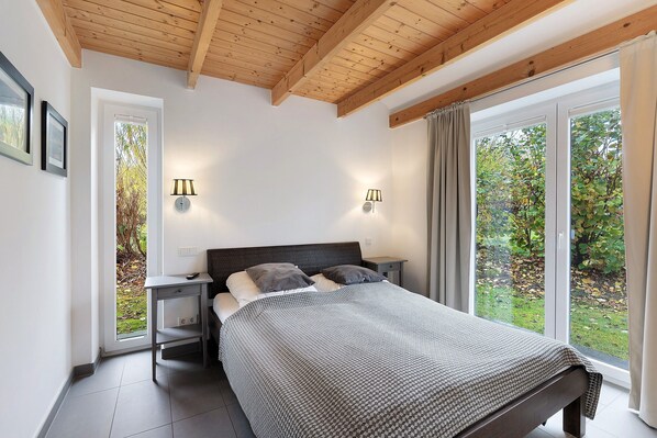 A bedroom with a wooden ceiling, a bed with a patterned bedspread, bedside lamps, a nightstand, and a window with a view of trees.