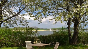 A grassy area with a table and two chairs, a tree, and a view of a lake with a sailboat.