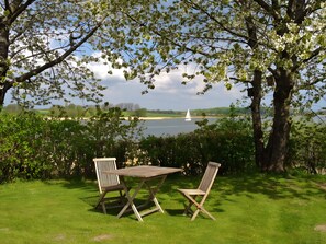 Een gazon met een tafel, twee stoelen, een boom en uitzicht op een meer met een zeilboot.
