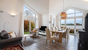 A spacious living area with a dining table, sofas, and a TV. Large windows offer views of the outdoors. - Holiday Home 'Schwanenuferhaus' with Sea View, Private Terrace and Wi-Fi (Kosel)