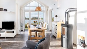 A modern living room with a TV, sofa, dining table, and chairs, a kitchen with a stove, and large windows overlooking a body of water. - Holiday Home 'Sonnenuferhaus' with Sea View, Private Terrace and Wi-Fi (Kosel)
