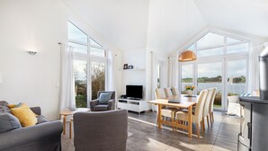 A spacious living area with a sofa, TV, dining table, and chairs, large windows with a view of a lake. - Holiday Home 'Sonnenuferhaus' with Sea View, Private Terrace and Wi-Fi (Kosel)