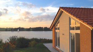A house with a tiled roof, large windows, and a view of a lake with a sailboat. - Holiday Home 'Sonnenuferhaus' with Sea View, Private Terrace and Wi-Fi (Kosel)