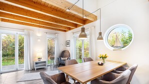 Un espace de salle à manger moderne avec une table en bois, des chaises grises, des luminaires suspendus et une vue sur la verdure à l’extérieur.
