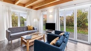 Un salon moderne avec un plafond en bois, un canapé gris, un canapé bleu, une table basse, un téléviseur et de grandes fenêtres donnant sur la forêt.