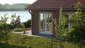 A house with a red-tiled roof, a glass door leading to a patio, and a view of a lake. - Holiday Home 'Schilfuferhaus' with Sea View, Private Terrace and Wi-Fi (Kosel)