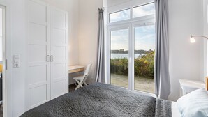 A bedroom with a large bed, a desk, and a view of a body of water through a sliding glass door. - Holiday Home 'Schilfuferhaus' with Sea View, Private Terrace and Wi-Fi (Kosel)