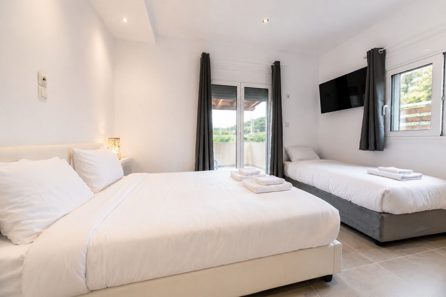 Habitación de hotel con una cama grande, una cama más pequeña, un televisor montado en la pared y un balcón con cortinas.