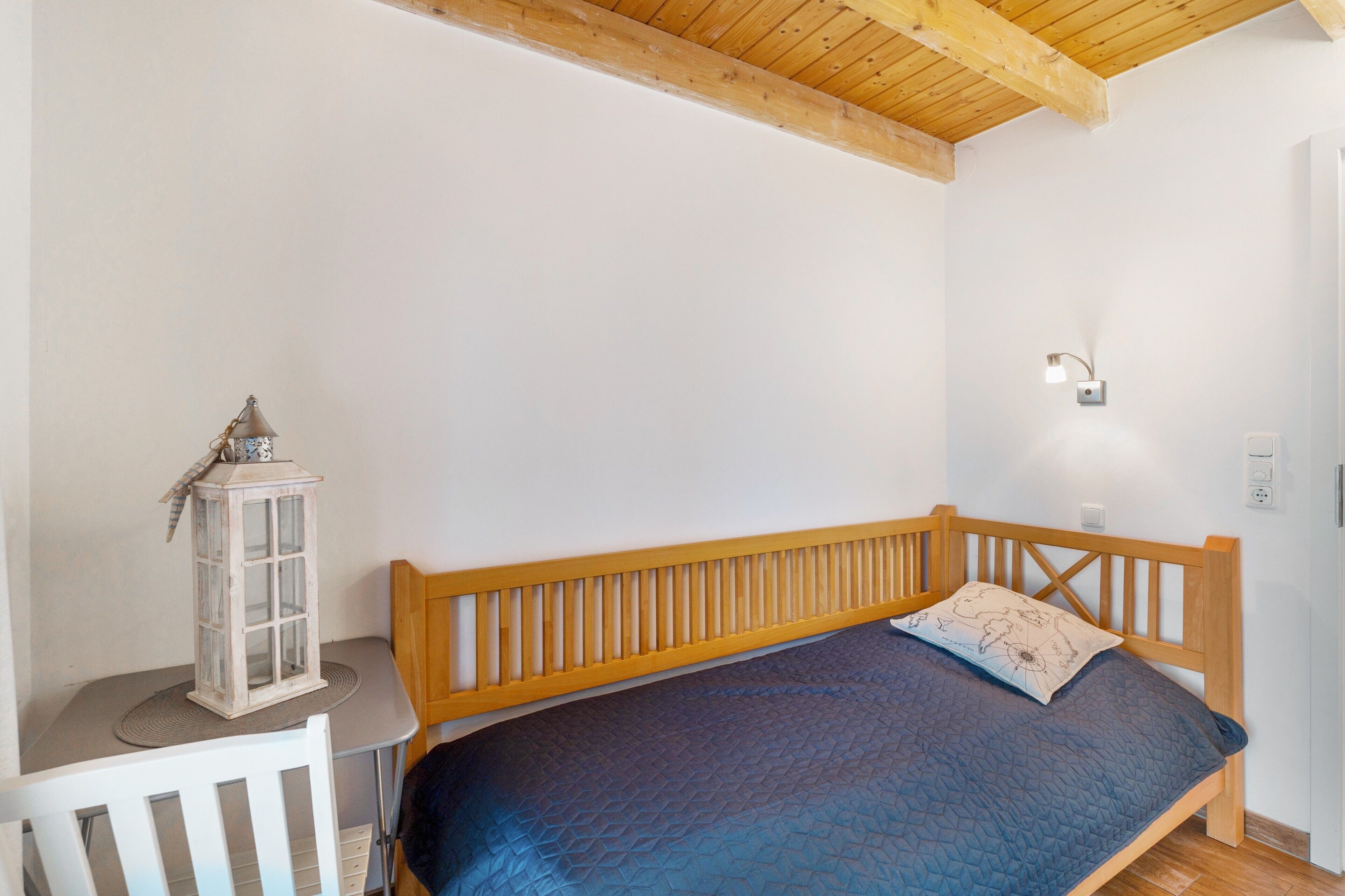 A bedroom with a wooden bed, a white chair, a small table, a wall-mounted light, and a decorative lantern.