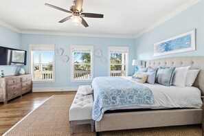 A bedroom with a bed, a ceiling fan, a dresser, and windows with a view of the outdoors.