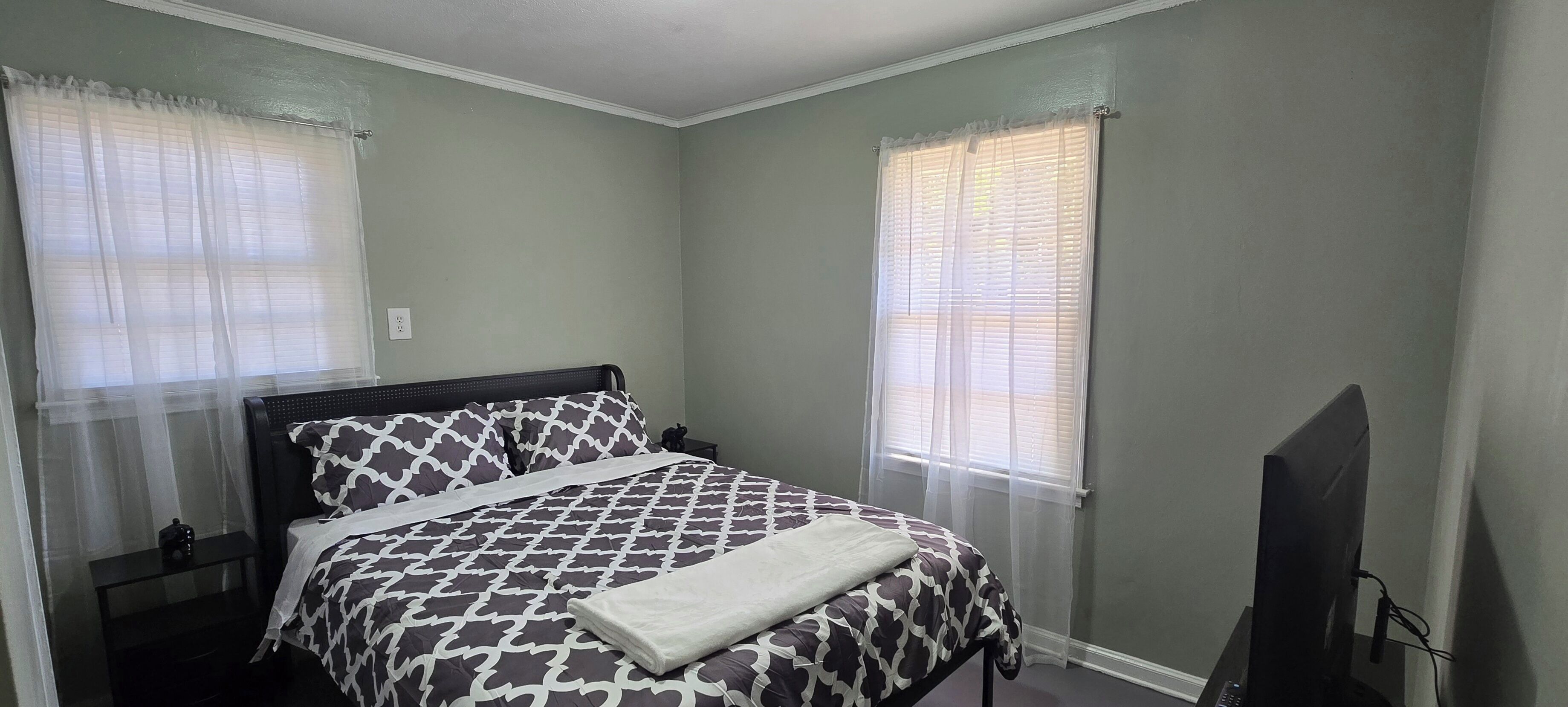 Um quarto com cama, mesa de cabeceira, televisão e duas janelas com cortinas leves.