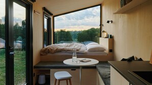 A compact bedroom with a bed, bedside table, and a window overlooking a garden. - Raus | Cabins am See mit Sauna + Hot-Tub (Lenzen)